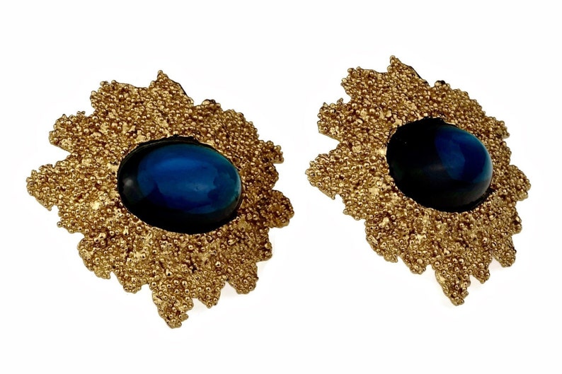 May include: A pair of gold earrings with a blue gemstone in the center of each earring. The earrings are shaped like a flower with a textured surface.