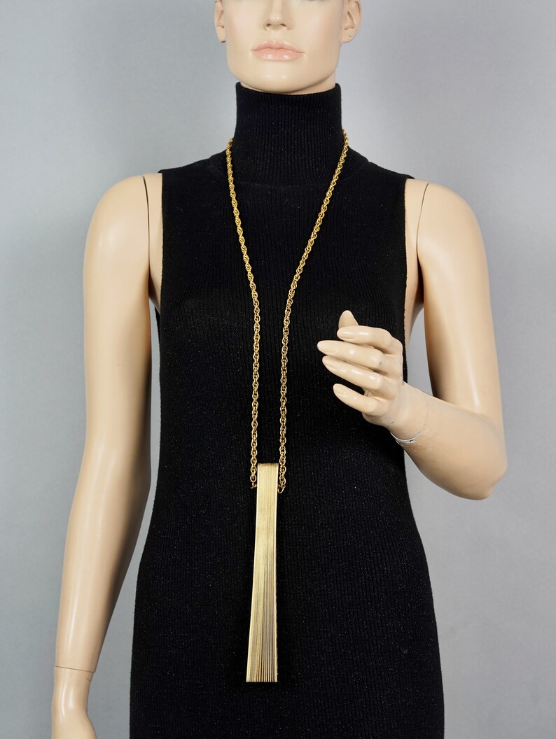 May include: A gold chain necklace with a long, rectangular pendant made of thin, vertical gold bars.