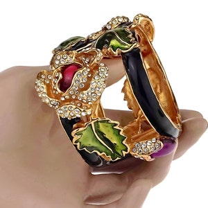 Puede incluir: Un brazalete de oro con esmalte negro y hojas y flores de esmalte verde y rojo. El brazalete est&aacute; adornado con diamantes de imitaci&oacute;n transparentes y una piedra cabuj&oacute;n p&uacute;rpura.