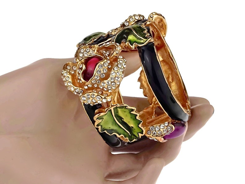Puede incluir: Un brazalete de oro con esmalte negro y hojas y flores de esmalte verde y rojo. El brazalete est&aacute; adornado con diamantes de imitaci&oacute;n transparentes y una piedra cabuj&oacute;n p&uacute;rpura.