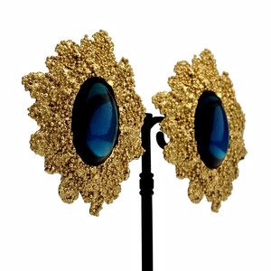 May include: A pair of gold tone earrings with a blue oval-shaped stone set in each. The earrings have a textured, floral design.