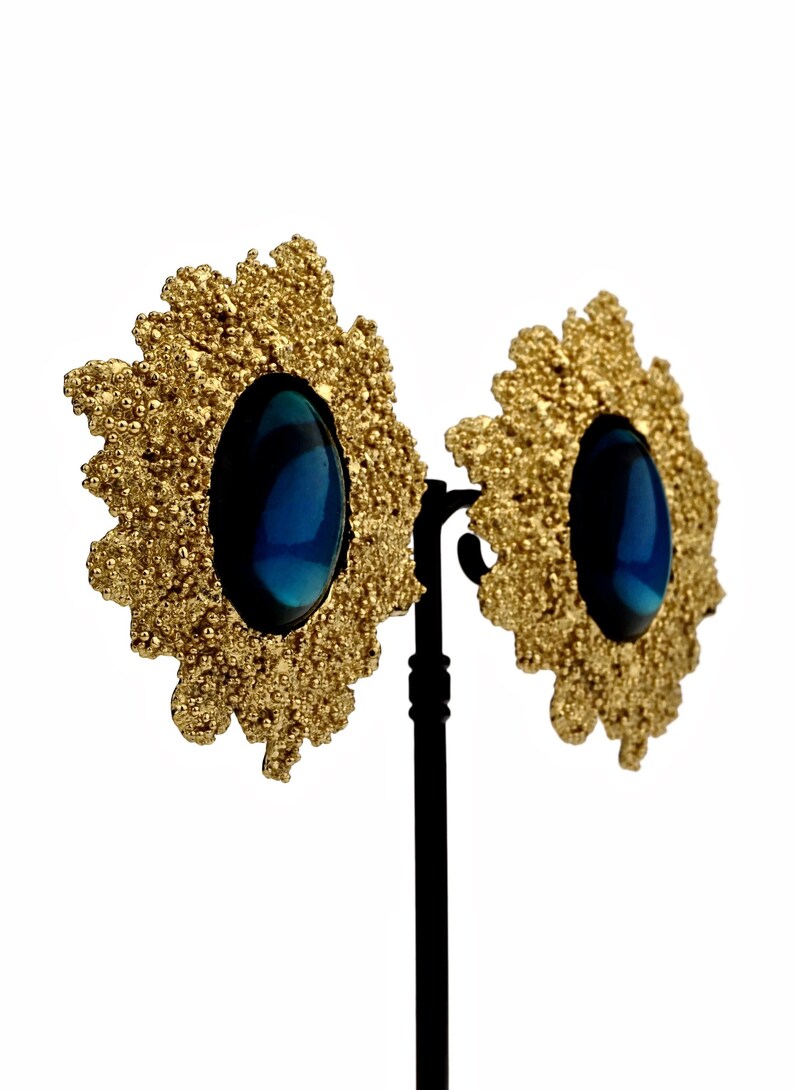 May include: A pair of gold tone earrings with a blue oval-shaped stone set in each. The earrings have a textured, floral design.