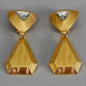 May include: A pair of gold tone dangle earrings with a clear crystal in the center of each earring. The earrings are shaped like a pyramid with a flat top.