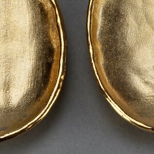 May include: A pair of gold-tone oval-shaped earrings with a textured surface. The earrings are marked with the word "Valentino" on the back.
