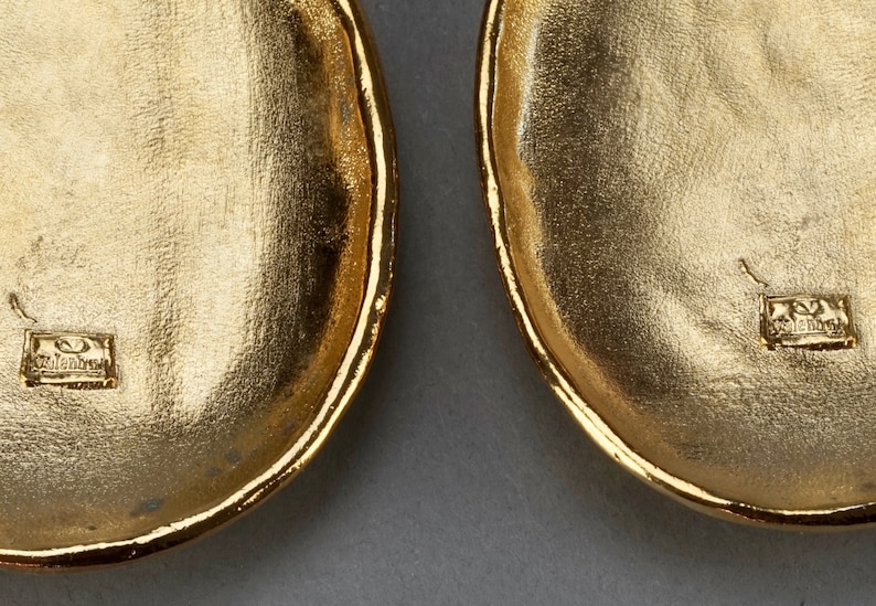 May include: A pair of gold-tone oval-shaped earrings with a textured surface. The earrings are marked with the word "Valentino" on the back.