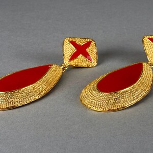May include: A pair of gold tone earrings with red enamel accents. The earrings are teardrop shaped and have a textured surface. The top of each earring features a square with a red cross.