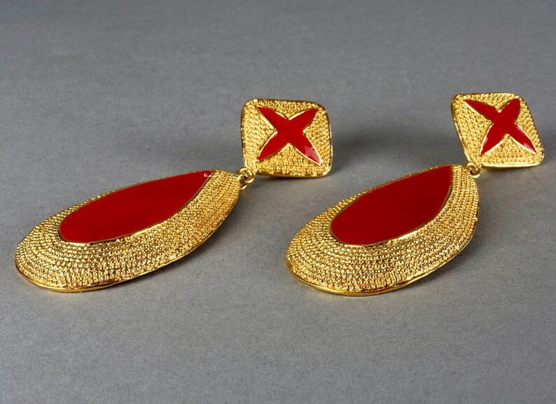 May include: A pair of gold tone earrings with red enamel accents. The earrings are teardrop shaped and have a textured surface. The top of each earring features a square with a red cross.