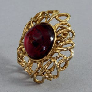 Könnte beinhalten: Ein goldfarbener Ring mit einem großen, ovalen, tiefroten Edelstein. Der Ring hat ein verziertes, offenes Design mit wirbelnden Formen. Der Edelstein hat eine glänzende Oberfläche, und der Ring ist vor einem neutralen Hintergrund platziert.