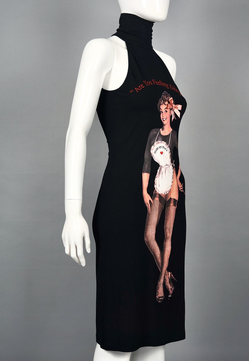 May include: Black halter dress with a pin-up graphic. The dress features a high neckline and a fitted silhouette. The graphic includes a vintage illustration of a woman in an apron, fishnet stockings, and the text "Are You Feeling Dirty?"