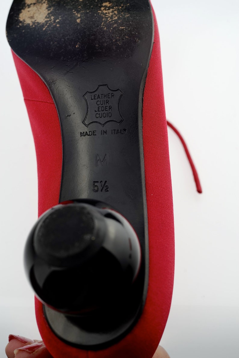 May include: Red leather heels with a black heel and a black ball on the bottom. The heel has the text "Leather Cuir Leder Cuoio Made in Italy" and the size "5 1/2" printed on it.