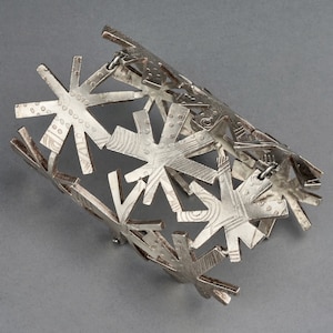 May include: Silver bracelet with a geometric design featuring multiple starburst shapes. The bracelet is made of metal and has a textured finish.