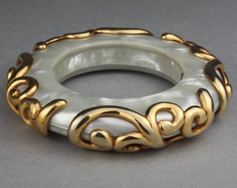 May include: A white bangle bracelet with a gold filigree design. The bracelet is made of a smooth, white material and has a delicate, intricate design.
