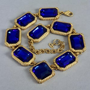 Puede incluir: Un collar dorado con grandes piedras preciosas rectangulares de color azul oscuro. Las piedras están engastadas en marcos dorados ornamentados y conectadas por una cadena dorada. El collar tiene un cierre de broche y un pequeño dije con las letras "CD".