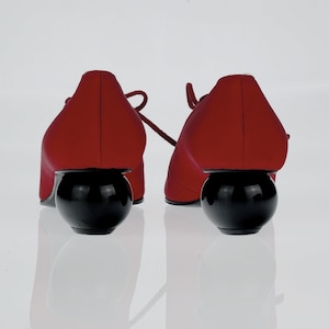 May include: A pair of red satin shoes with black ball heels. The shoes have laces and are sitting on a white surface.