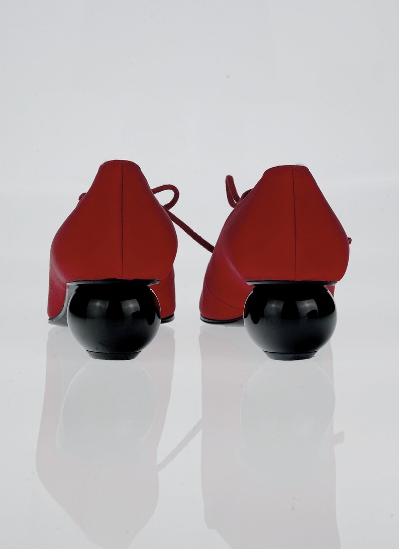 May include: A pair of red satin shoes with black ball heels. The shoes have laces and are sitting on a white surface.