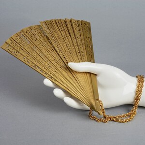 May include: A gold-colored hand fan with intricate cutouts and a gold chain attached. The fan is held in a white mannequin hand.