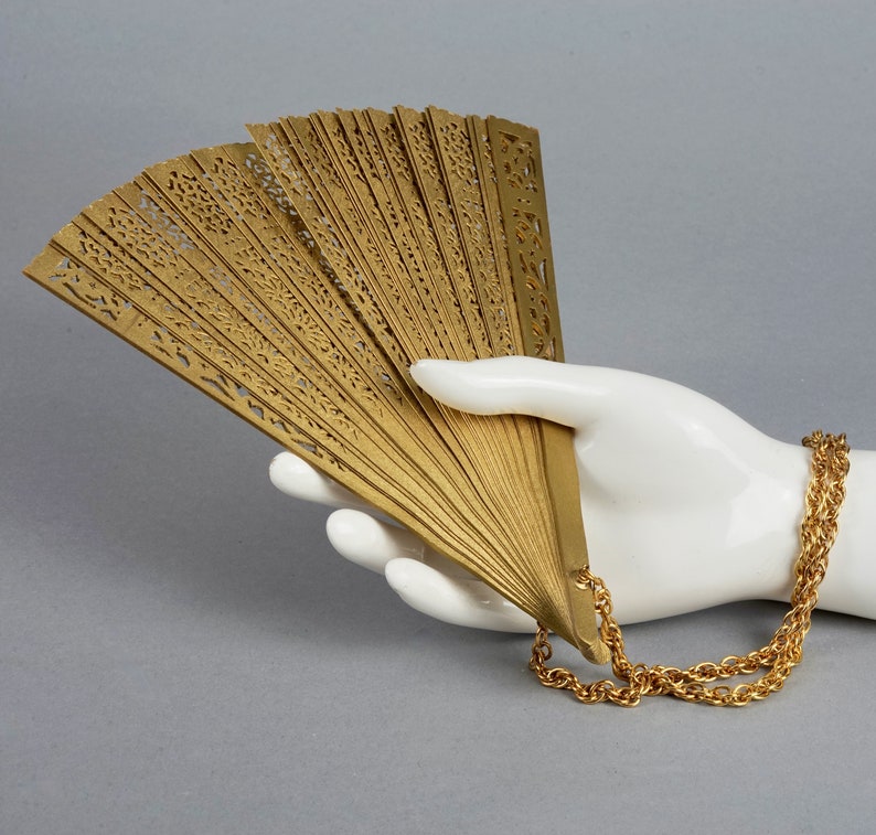 May include: A gold-colored hand fan with intricate cutouts and a gold chain attached. The fan is held in a white mannequin hand.