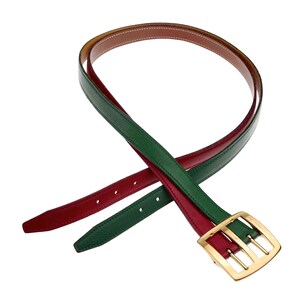 Pode incluir: Um cinto de couro revers&iacute;vel com um design verde e vermelho. O cinto tem uma fivela dourada com tr&ecirc;s barras verticais.