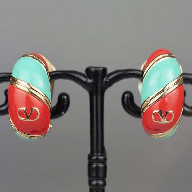 May include: A pair of red and turquoise hoop earrings with a gold logo. The earrings are on a black stand.