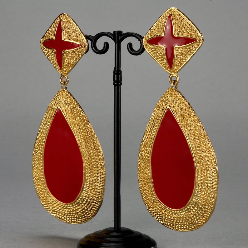 May include: A pair of gold tone dangle earrings with red enamel teardrop shapes. The earrings feature a textured gold finish and a red cross design at the top.