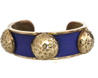 Op de afbeelding: Een goudkleurige manchetarmband met een getextureerde blauwe middenband. De armband heeft drie grote, getextureerde, goudkleurige cirkelvormige accenten. Het open manchetontwerp is zichtbaar.