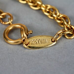May include: A gold chain necklace with a clasp and a small oval tag that says "MOSCHINO" in all caps.