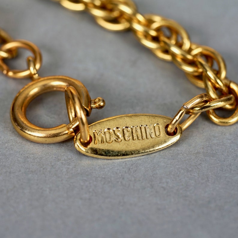 May include: A gold chain necklace with a clasp and a small oval tag that says "MOSCHINO" in all caps.