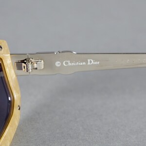 May include: Close-up of the arm of a pair of beige sunglasses with a silver metal frame. The sunglasses have a Christian Dior logo on the arm.