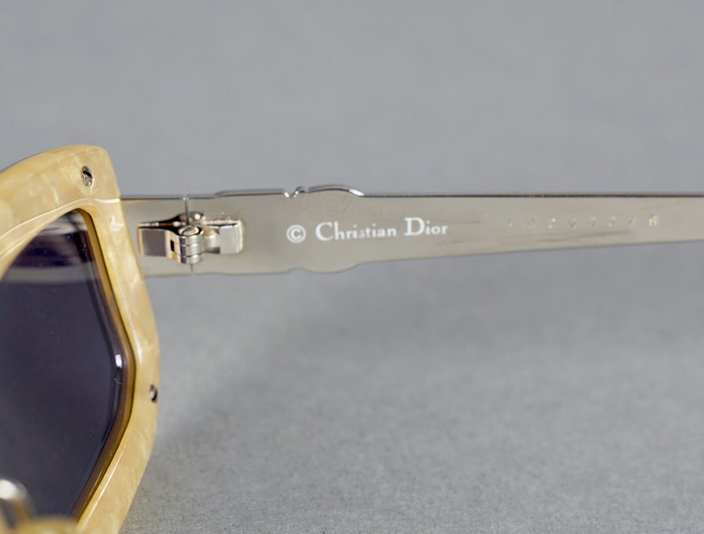 May include: Close-up of the arm of a pair of beige sunglasses with a silver metal frame. The sunglasses have a Christian Dior logo on the arm.