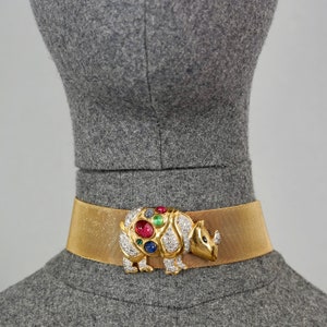 May include: A gold mesh choker necklace with a large, jeweled rhinoceros charm. The charm is set with clear, red, green, and blue gemstones.