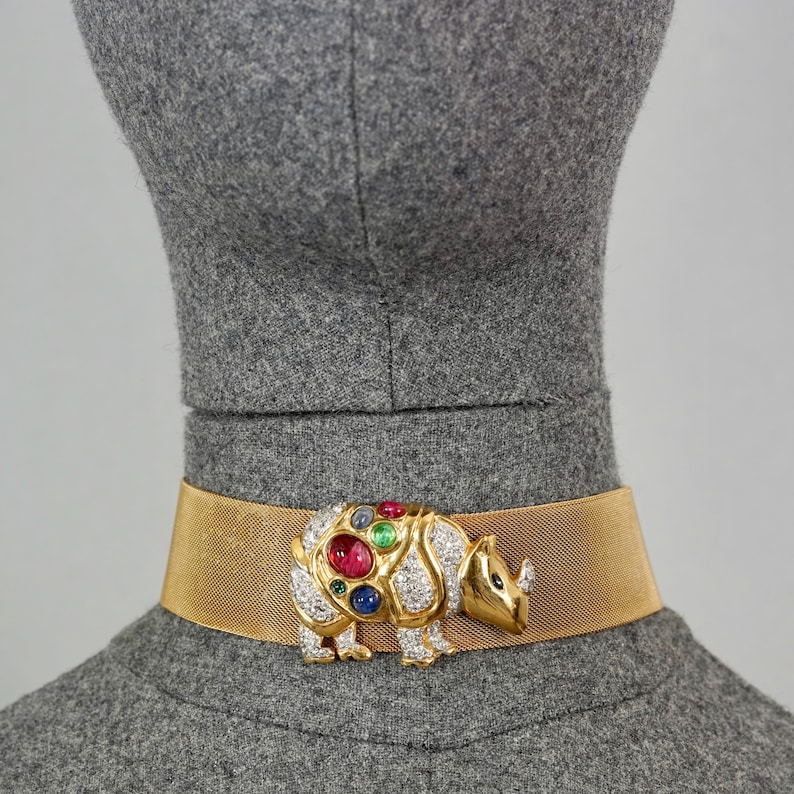 May include: A gold mesh choker necklace with a large, jeweled rhinoceros charm. The charm is set with clear, red, green, and blue gemstones.