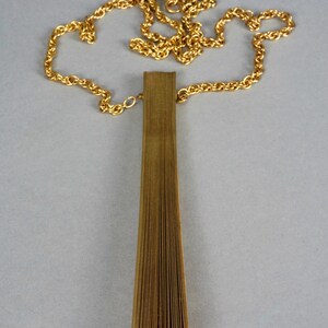 May include: A gold chain necklace with a long, thin, gold pendant that is folded like a fan.