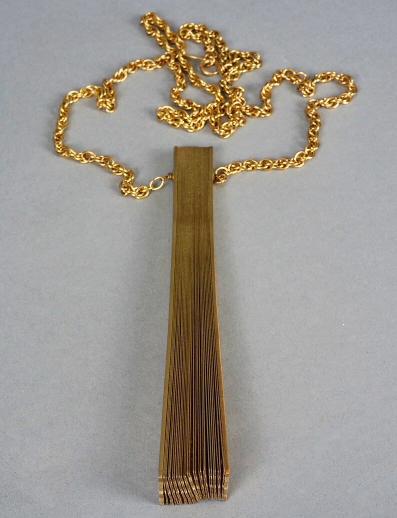 May include: A gold chain necklace with a long, thin, gold pendant that is folded like a fan.
