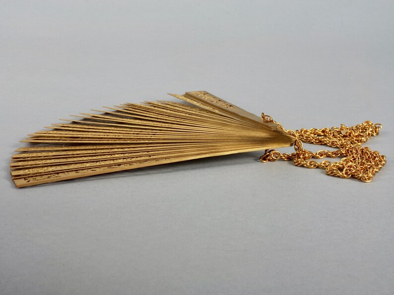 May include: A gold-colored fan-shaped pendant necklace with a chain. The fan is made of multiple thin metal pieces that are connected together.