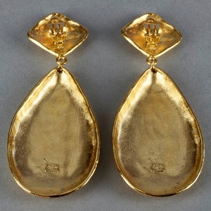 May include: A pair of gold-tone teardrop-shaped earrings with a square-shaped top. The earrings have a textured surface and a shiny finish.
