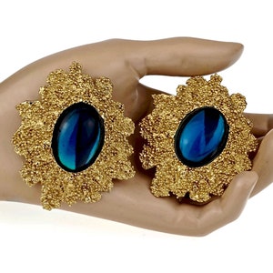 May include: A pair of large, gold-tone statement earrings with blue, oval-shaped stones set in the center. The earrings have a textured, floral design.