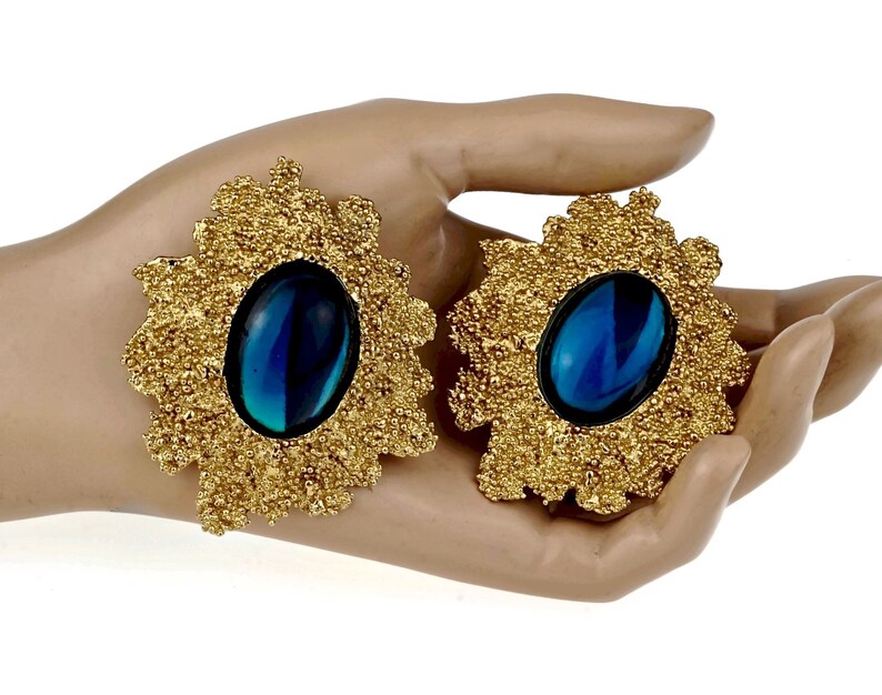 May include: A pair of large, gold-tone statement earrings with blue, oval-shaped stones set in the center. The earrings have a textured, floral design.