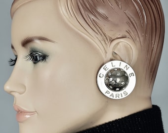 Op de afbeelding: Zilverkleurige clip-on oorbellen met een rond ontwerp. De oorbellen hebben de tekst "CELINE PARIS" en een getextureerd midden met strass.