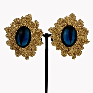 May include: A pair of gold tone clip-on earrings with a blue oval-shaped stone in the center of each earring. The earrings are in the shape of a flower with a textured design.