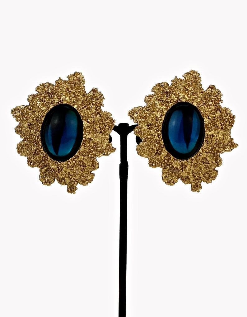 May include: A pair of gold tone clip-on earrings with a blue oval-shaped stone in the center of each earring. The earrings are in the shape of a flower with a textured design.