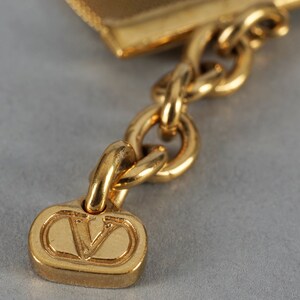 May include: A gold-toned chain with a V-shaped charm. The charm is attached to a chain link and has a raised V design.