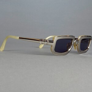 May include: Silver-toned sunglasses with a rectangular frame and dark lenses. The arms of the sunglasses are made of a cream-colored material.