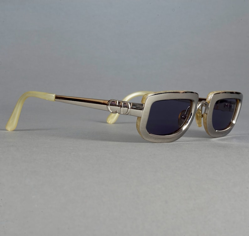 May include: Silver-toned sunglasses with a rectangular frame and dark lenses. The arms of the sunglasses are made of a cream-colored material.