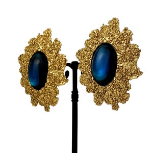 May include: A pair of gold tone earrings with blue oval-shaped stones set in a textured, flower-shaped design.