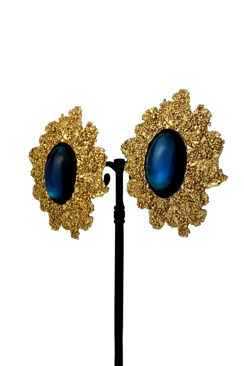 May include: A pair of gold tone earrings with blue oval-shaped stones set in a textured, flower-shaped design.