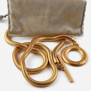 Puede incluir: Un collar de cadena dorado con un gran colgante redondo que dice "Paco Rabanne Paris". La cadena es de estilo serpiente y el colgante tiene un anillo en el centro.