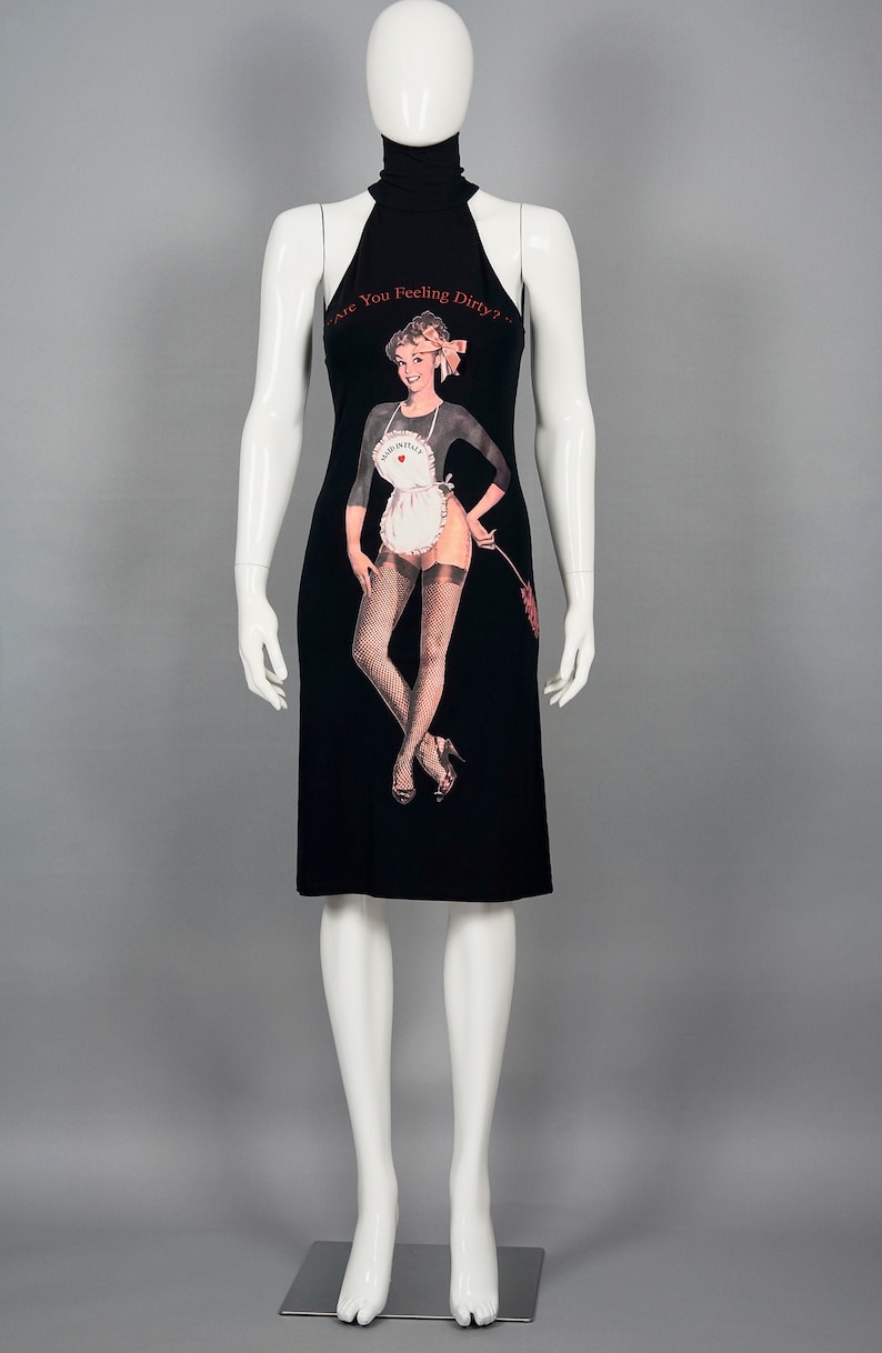 May include: Black halter dress with a pin-up graphic. The dress features a high neckline and a knee-length hem. The graphic includes the text "Are You Feeling Dirty?" and an image of a woman in a maid outfit. The dress is displayed on a mannequin.