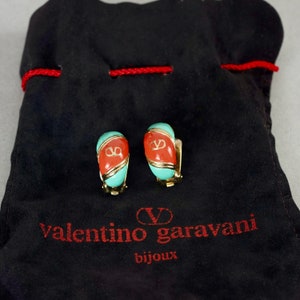 May include: A pair of gold-tone clip-on earrings with red and turquoise enamel. The earrings feature the Valentino Garavani logo in gold. The earrings are displayed on a black velvet pouch with the Valentino Garavani logo in red.