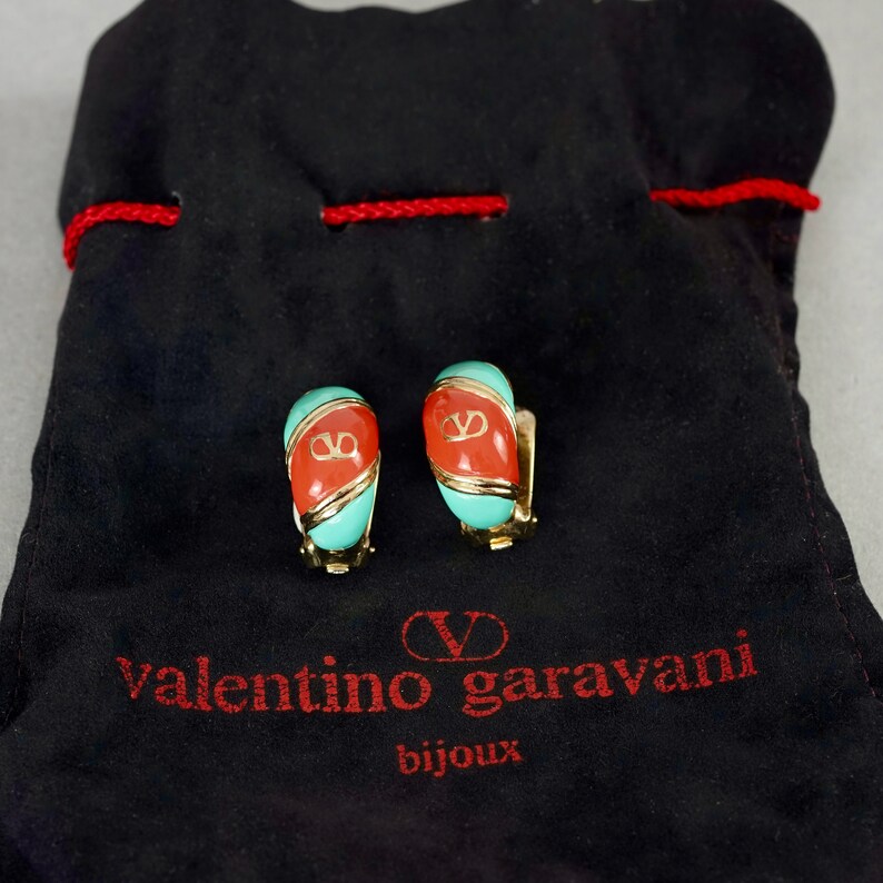 May include: A pair of gold-tone clip-on earrings with red and turquoise enamel. The earrings feature the Valentino Garavani logo in gold. The earrings are displayed on a black velvet pouch with the Valentino Garavani logo in red.