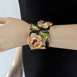 Puede incluir: Un brazalete de tono dorado con esmalte negro y pedrer&iacute;a. El brazalete presenta un dise&ntilde;o floral con hojas verdes y un centro rojo.